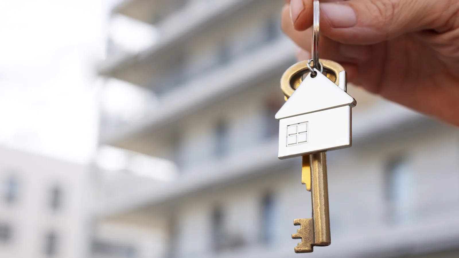 Main tenant un porte-clés en forme de maison devant un immeuble résidentiel, symbole d’un investissement locatif